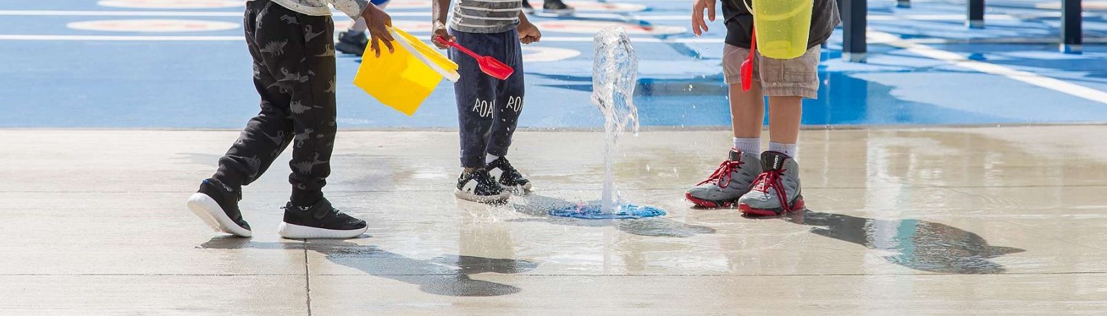 Three children's legs and feet can be seen playing in a water splash area with a plastic bucket.