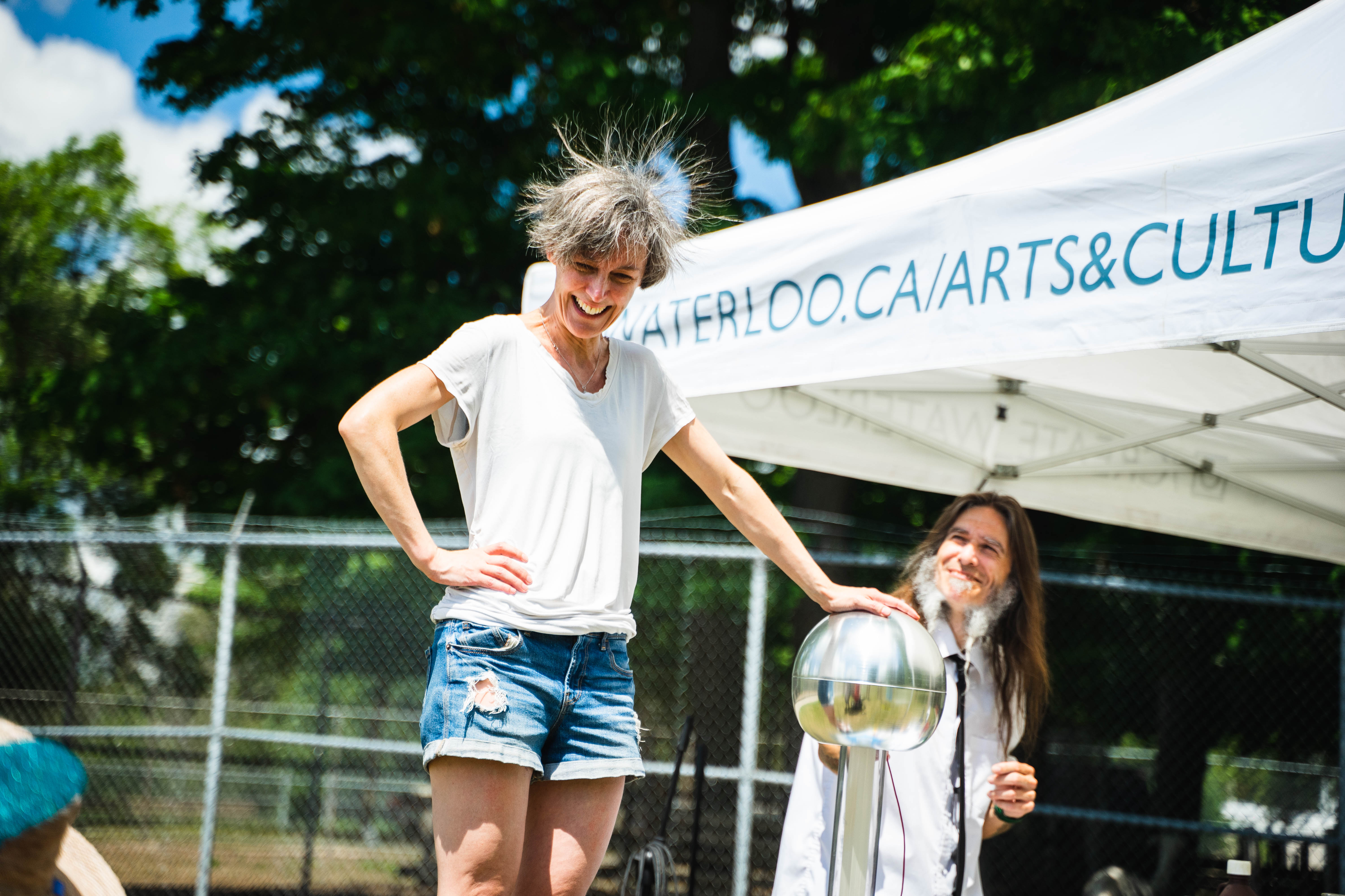 A person stands laughing on a platform touching a Van de Graaff generator, causing their hair to static while another person stands and smiles nearby at an outdoor event.