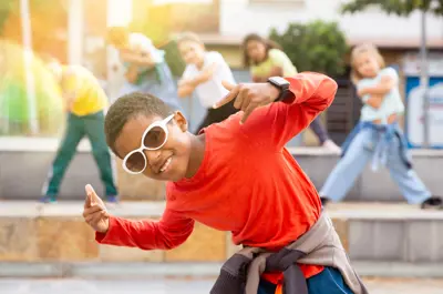 A person wearing a red shirt and white sunglasses poses energetically, with several others dancing in the background in an outdoor urban setting.