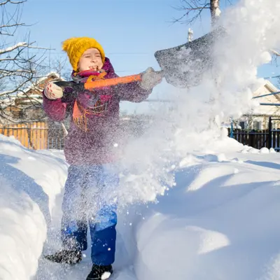 A child in winter clothing shovels snow along a path, tossing a large spray of snow into the air on a sunny day.