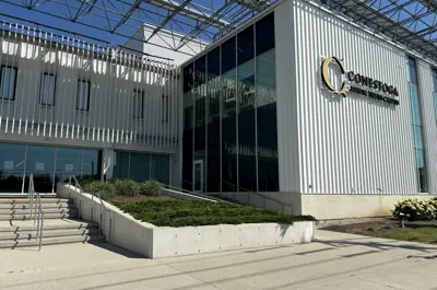 Conestoga College building exterior with modern white vertical panels, large glass windows, a metal canopy above, and steps leading to the main entrance on a sunny day.