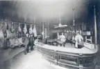 Black and white photo of the interior of Fischer's Meat Market. Two customers stand in front of counter while two butchers stand behind. Large cuts of meat cover the walls.