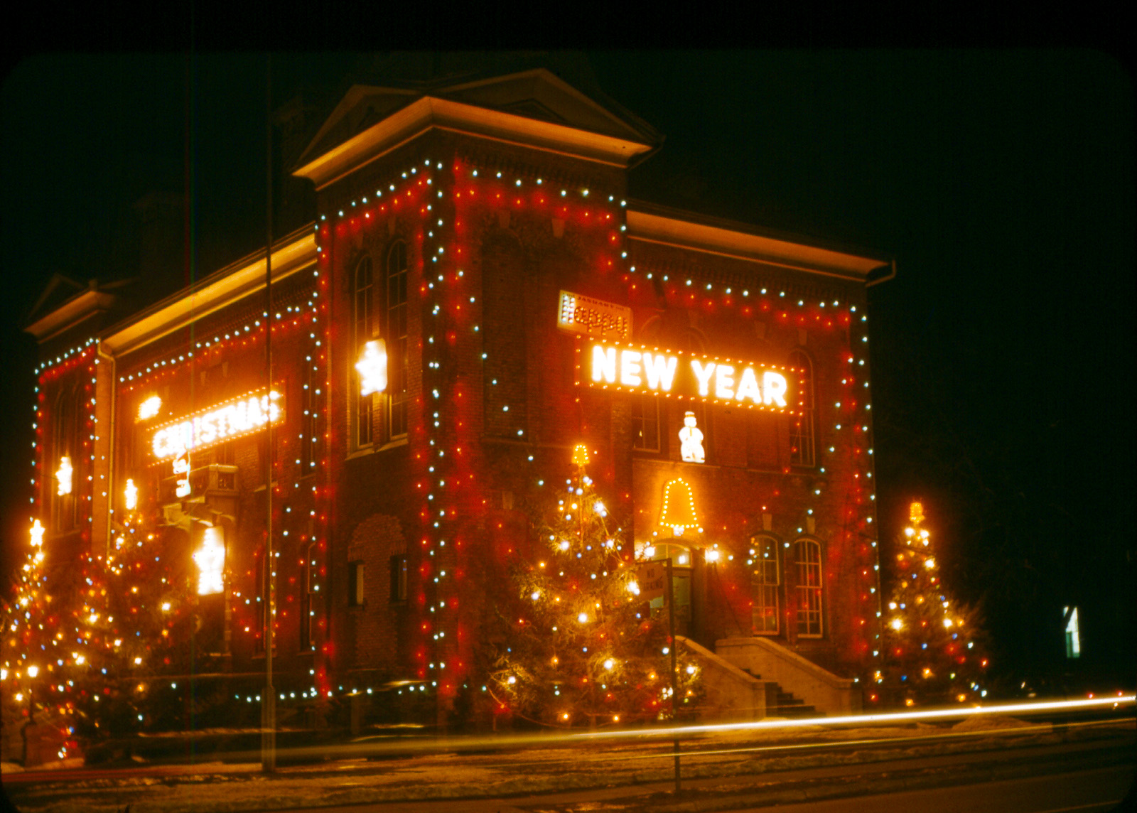 A brick building covered in colourful Christmas lights and decorations at night, with bright signs that read “Christmas” and “New Year,” and lit trees around the entrance