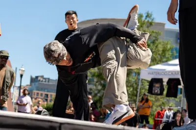 A person breakdancing on an outdoor stage, bending down with one leg raised and an audience of people energetically observing.