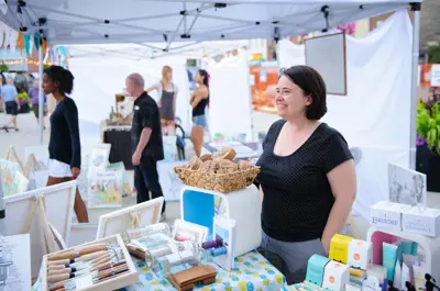 People stand under outdoor market booths, with various products and artworks displayed underneath. 