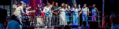Musicians and singers perform under stage lights at an outdoor concert, with a crowd watching.