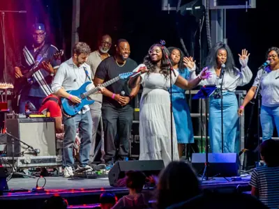 Musicians and singers perform under stage lights at an outdoor concert, with a crowd watching.