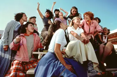 A group of people from the film Grease sit on bleachers, cheering and smiling under a clear, blue sky.