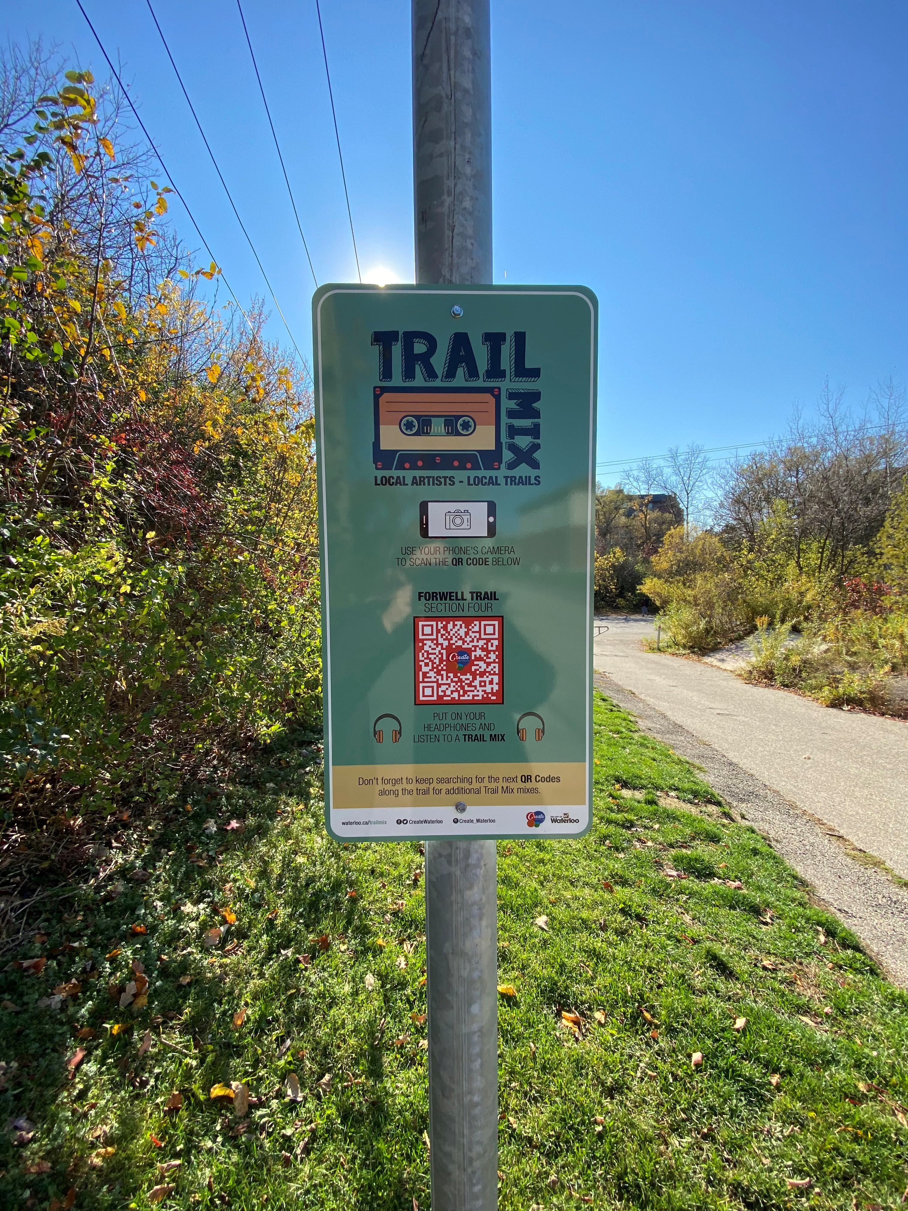 A Trail Mix sign with a QR code stands beside a walking trail, inviting people to scan and listen to local music.