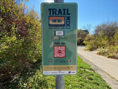 A Trail Mix sign with a QR code stands beside a walking trail, inviting people to scan and listen to local music.