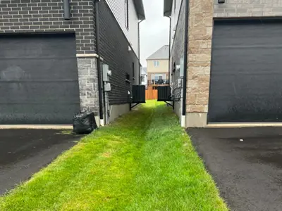 Image of a side-swale showing a shallow, grassed channel along a property line designed to direct stormwater runoff away from a house toward drainage infrastructure.