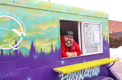 Person smiling out a purple and green food truck window on winters day that features a large logo of a white goose and text that says 'coffee roasters'.