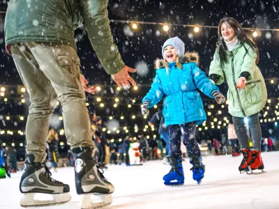 A child skates with two adults on an outdoor rink at night, smiling as snow falls.