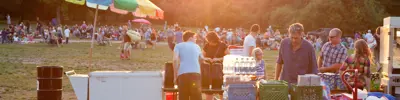 Outdoor community event in Waterloo park at sunset, featuring people browsing a food vendor booth with a crowd of people sitting on the grass in the background.