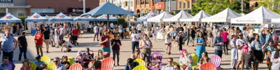 Crowd of people browsing outdoor vendor booths while others sit in colourful chairs in Waterloo Public Square.