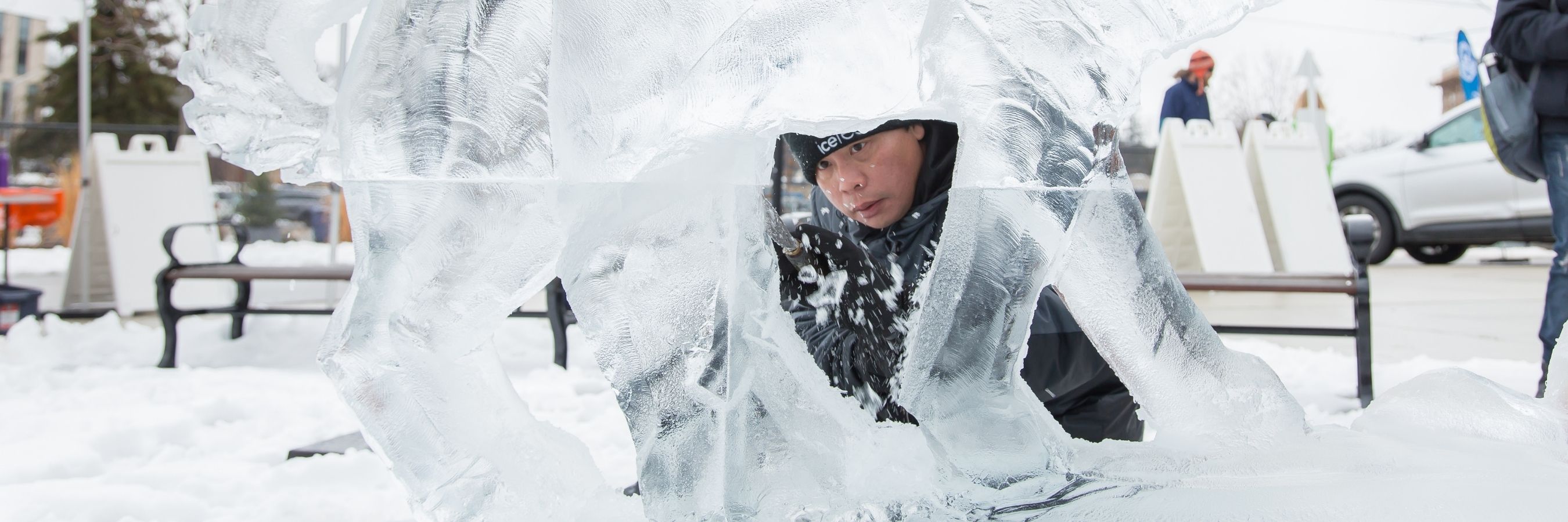 An artist carves a large ice sculpture outdoors in the snow.