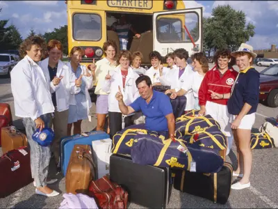 Women from the 1987 KW Civitan Ladies Softball team stand around luggage behind a school bus and hold up their index fingers.