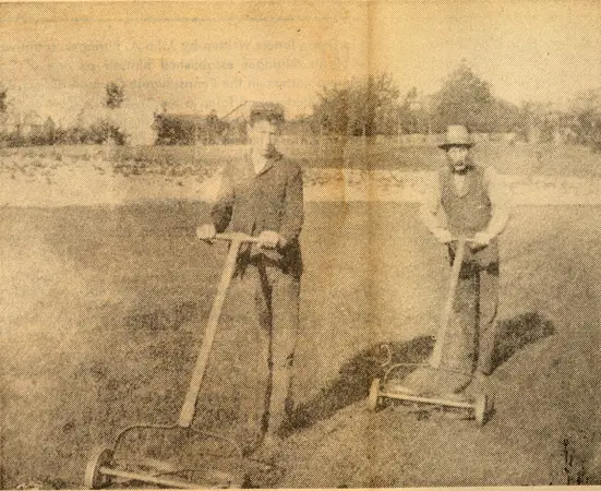 Sepia-toned photograph of park superintendent Andrew McIntyre and his young son standing and hand‑mowing grassy park grounds beside a lakeside vista, circa early 1900s.