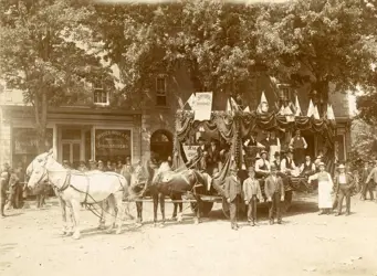 Black and white photo of horse-drawn parade float covered in fabric with men sitting on and standing around the float