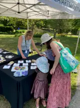 Museum volunteer at an outdoor event with a table showcasing current exhibits to a woman and child
