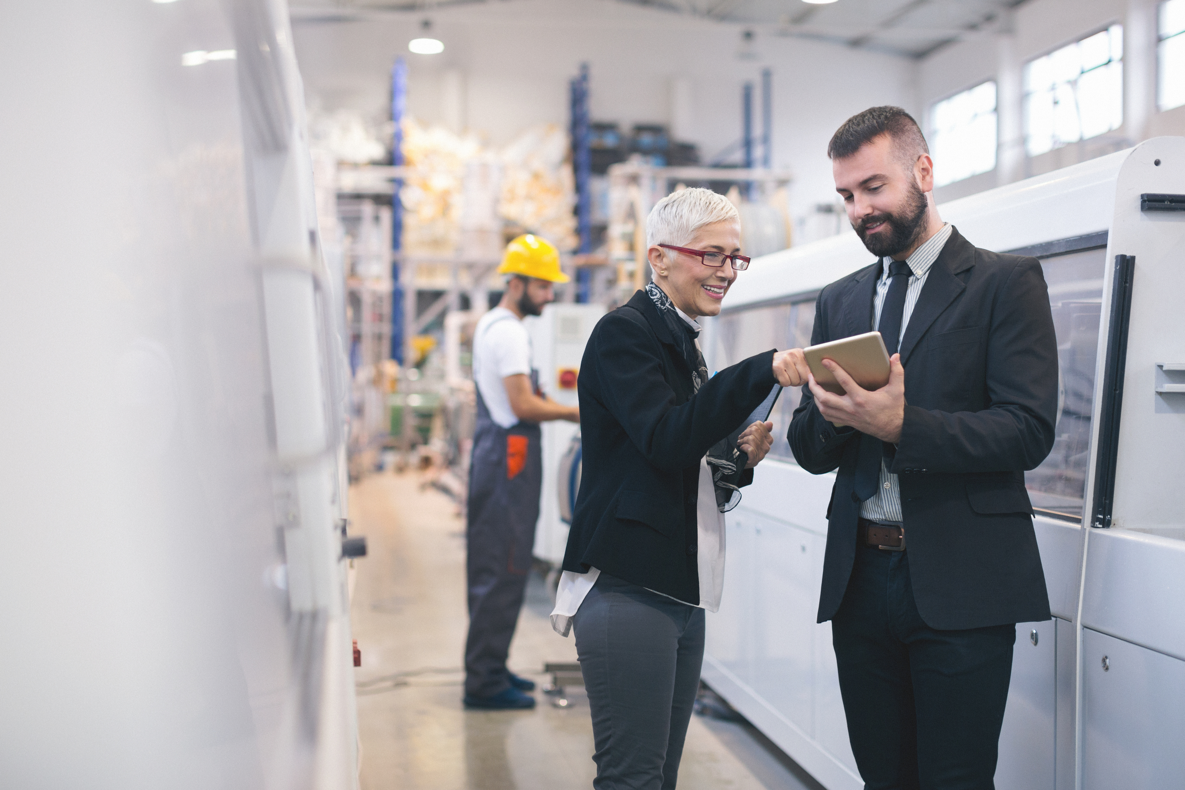 Two professionals stand in a modern manufacturing space, reviewing information on a tablet beside industrial equipment.