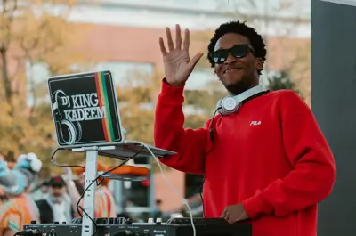 Person wearing a red sweater and headphones stands in front of DJ equipment and a laptop titled 'DJ King Kadeem' and waves at the camera.
