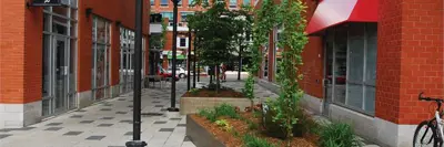 A narrow pedestrian walkway between two brick buildings with raised planters, small trees, outdoor seating and storefronts, creating a publicly accessible space.