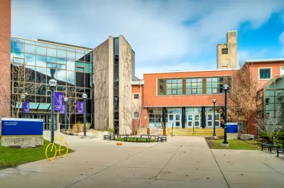 A courtyard on the Laurier campus with brick and glass buildings, banners, benches, and a yellow bike sculpture on a cloudy day.