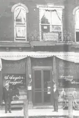 Black and white photo of storefront with two men in front.