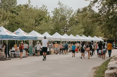 An outdoor event with people walking down a road along rows of white tents by green trees.