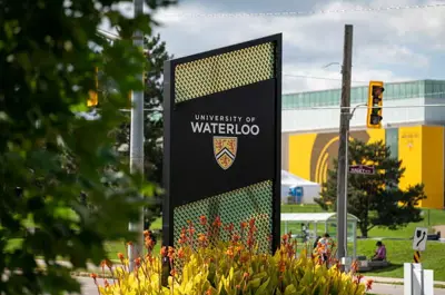 The University of Waterloo entrance sign stands in front of landscaped plants, with campus buildings and traffic lights in the background.