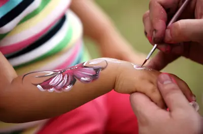 An arm being painted with a pink butterfly design. The background is blurred of a striped shirt and pink pants.