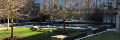 A landscaped courtyard with open lawn, young trees, circular seating areas and paved walkways, located between modern glass buildings.