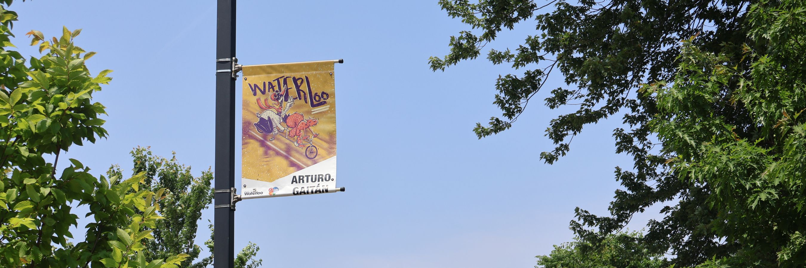A street banner with colourful artwork hangs from a pole on a sunny day, surrounded by trees.