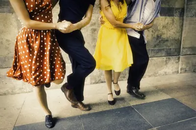 Four people are dancing closely together against a stone wall wearing colorful clothing.