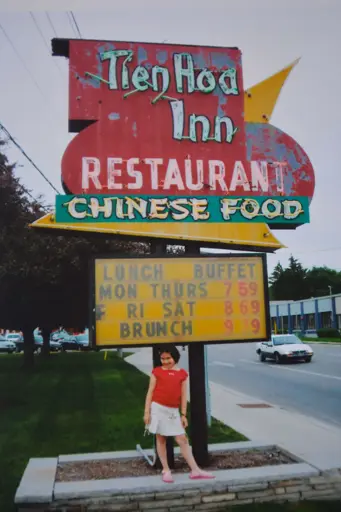 Image of restaurant sign, reads “Tien Hoa Inn Restaurant, Chinese Food” with child standing before it.