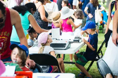 A lively outdoor community event in Waterloo Park with people of varying ages sitting and standing near tables using typewriters.