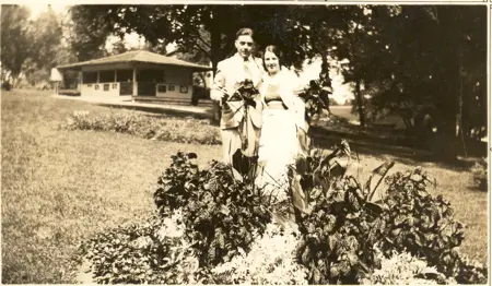 Black-and-white portrait of Clarence and Ruth Bohlender posing beside a flowerbed in Waterloo Park, with a concession stand visible in the background, circa early 1900s.