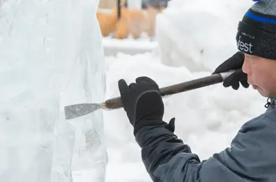 A person in warm winter clothing sculpts an ice block with a chisel in a snowy outdoor setting.