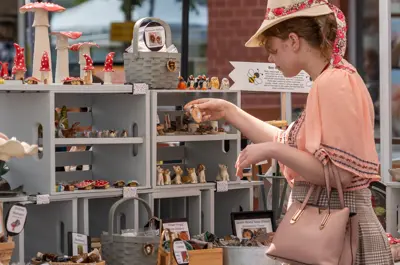 Person wearing skirt and hat stands under outdoor stall while interacting with a display of ceramic items such as garden gnomes and mushrooms.